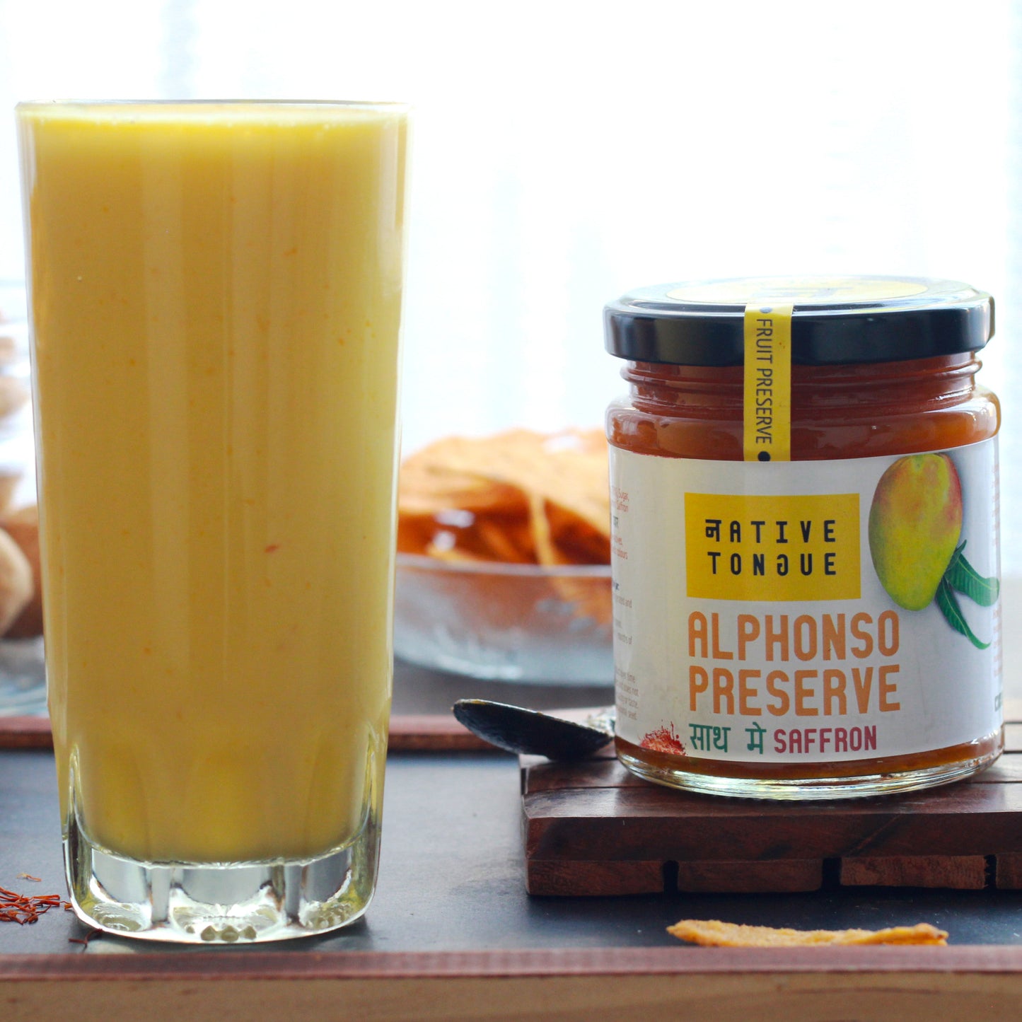 Two glasses of a Mango smoothie next to a jar of 'Alphonso Preserve' on a wooden tray.