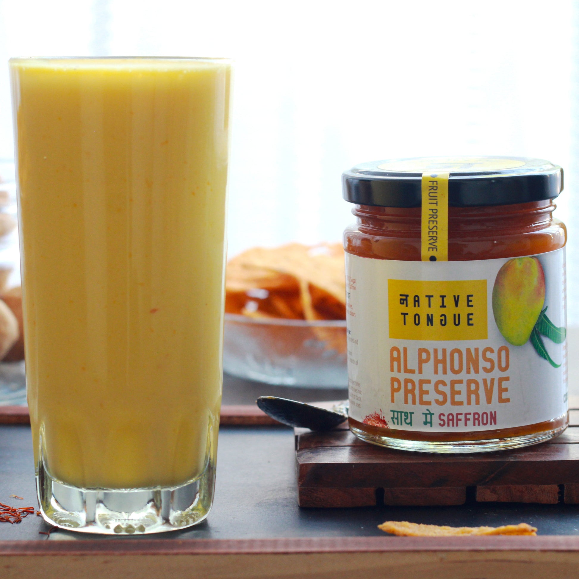 Two glasses of a Mango smoothie next to a jar of 'Alphonso Preserve' on a wooden tray.