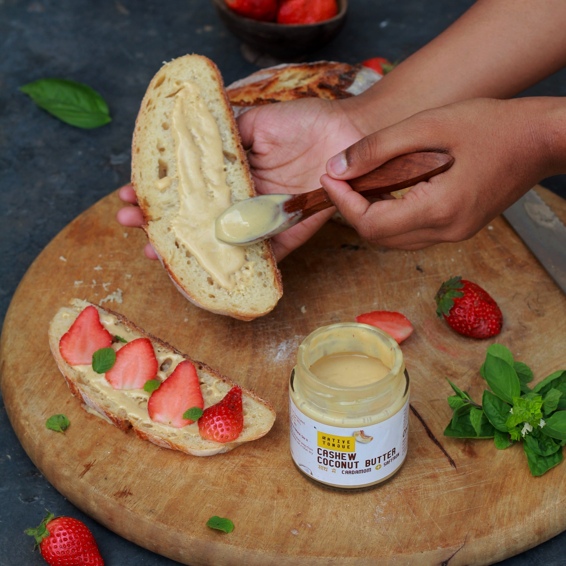 Person spreading cashew coconut butter on bread with strawberries on a wooden board.