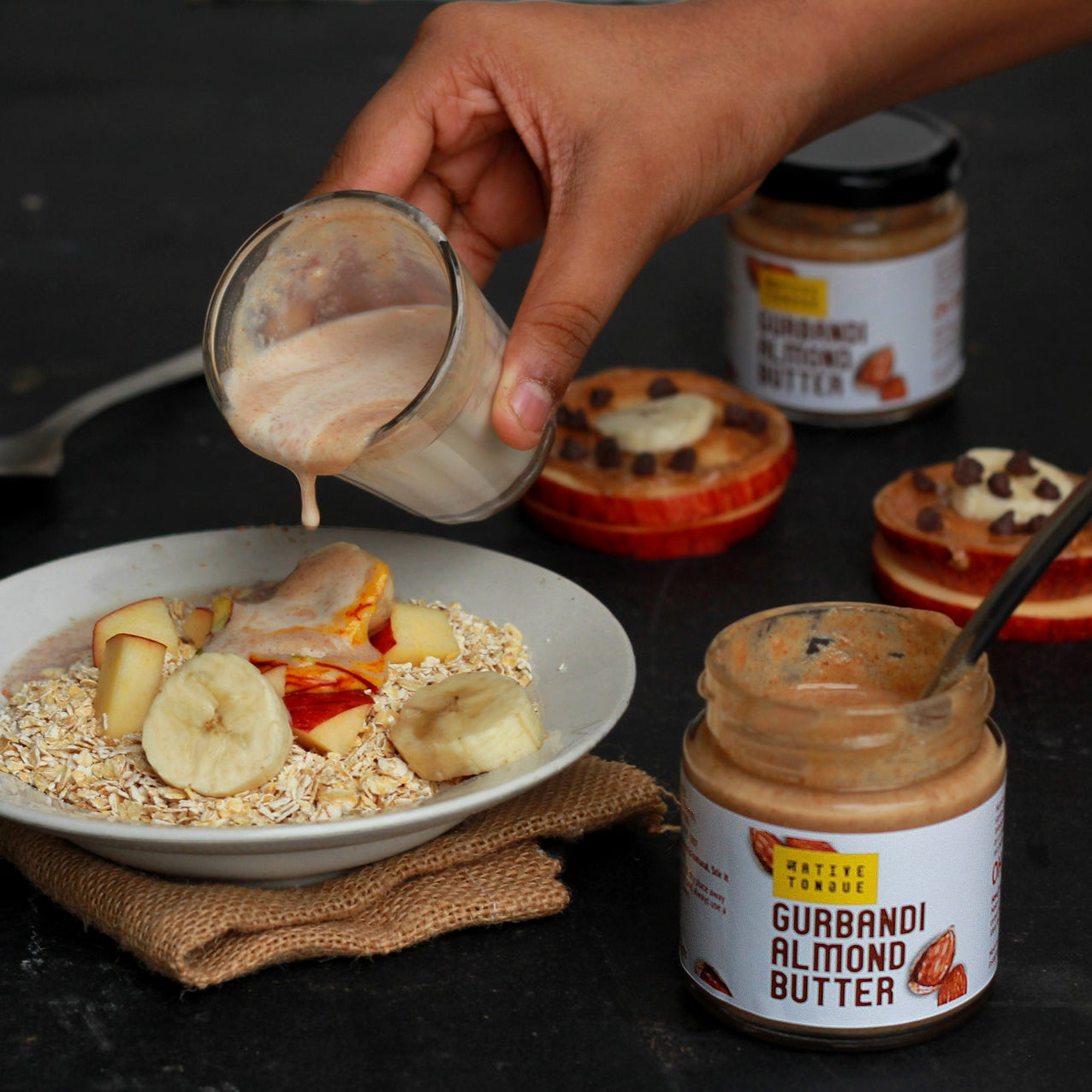 Person pouring almond butter onto a bowl of oatmeal with fruits, surrounded by jars of almond butter.