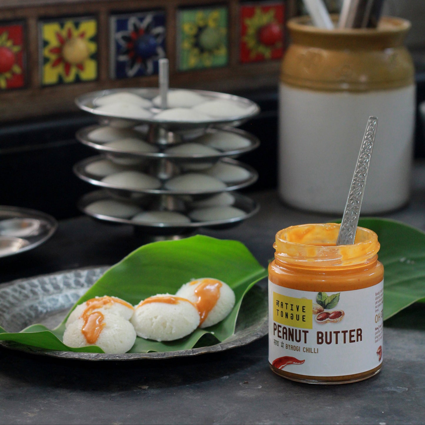 Jar of peanut butter with a spoon on a leaf, surrounded by kitchen items.