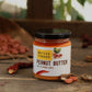 Jar of peanut butter with a visible brand label on a rustic wooden surface with peanuts and chili peppers.