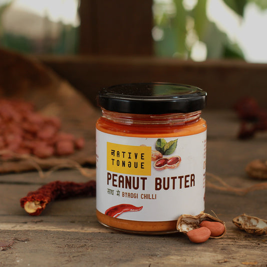 Jar of peanut butter with a visible brand label on a rustic wooden surface with peanuts and chili peppers.