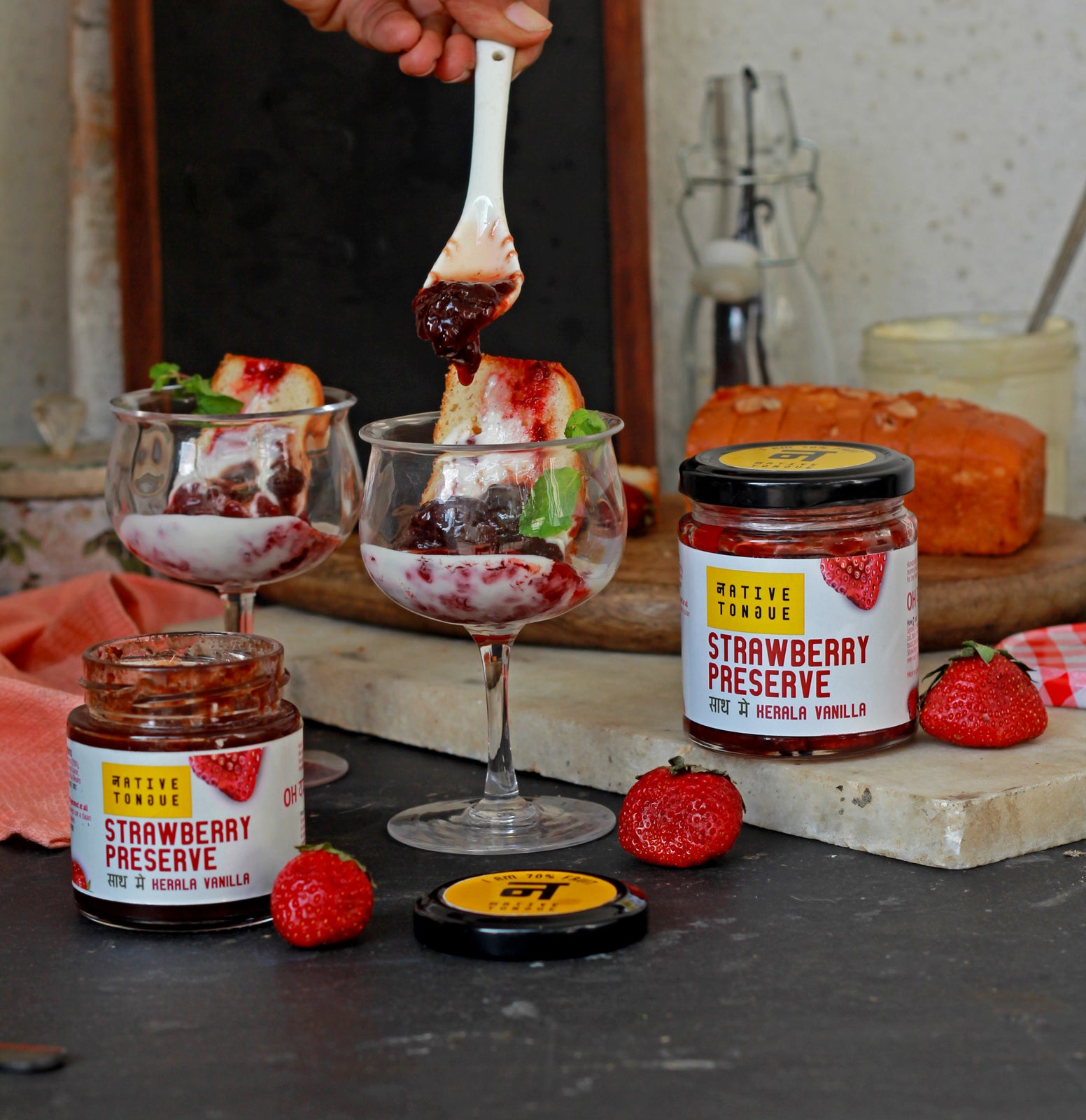 Strawberry preserve being spread on a dessert with jars and strawberries on a table.