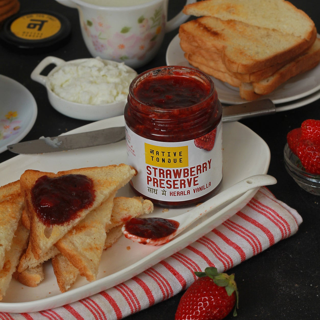 Jar of strawberry preserve on a plate with toast and a strawberry on a dark surface.