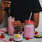 Person pouring a pink drink into a glass with a jar of strawberry preserve on a table.