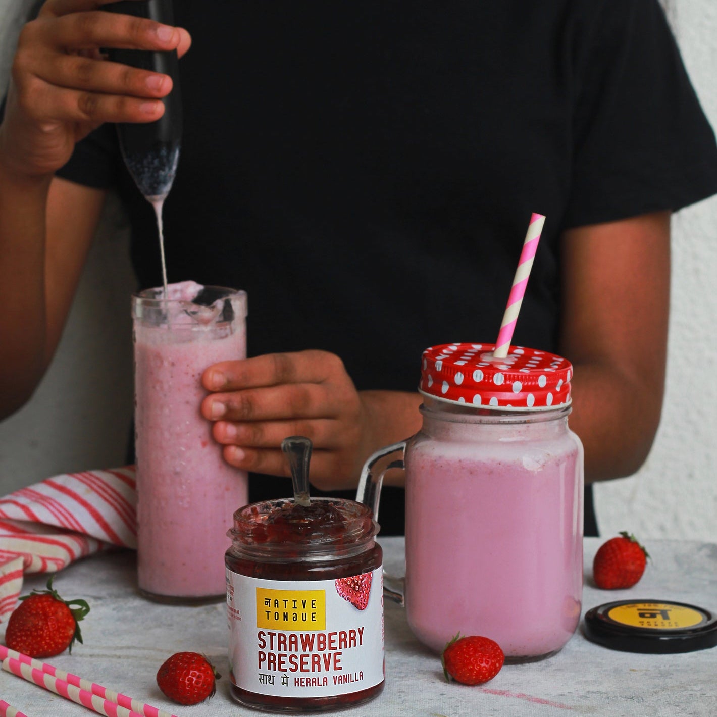 Person pouring a pink drink into a glass with a jar of strawberry preserve on a table.