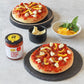 Two small pizzas with toppings on black stone boards, accompanied by a jar of sauce and small bowls of ingredients.