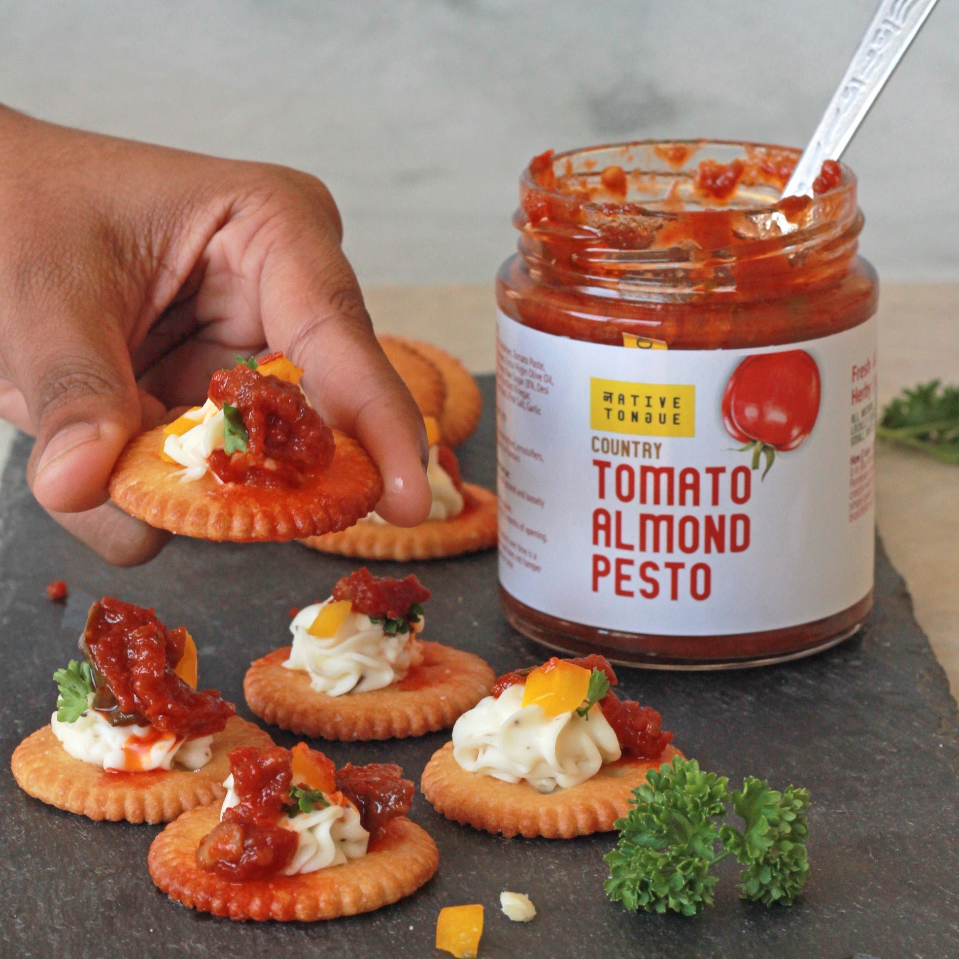 Jar of Tomato Almond Pesto with crackers being topped with the pesto.