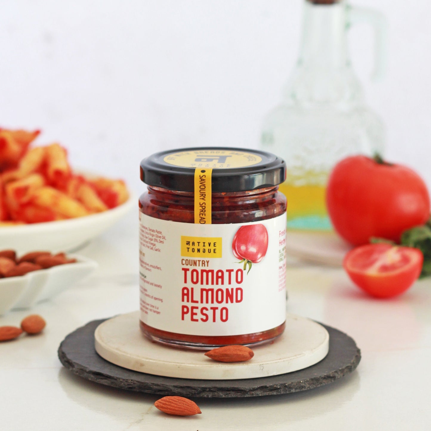 Jar of Tomato Almond Pesto on a white surface with ingredients in the background