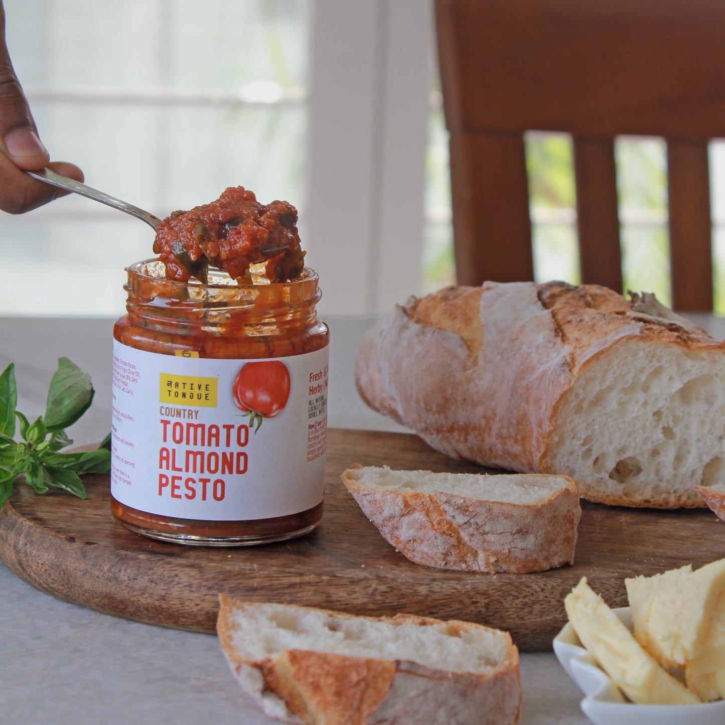 Jar of Tomato Almond Pesto on a wooden board with bread slices and a spoon.